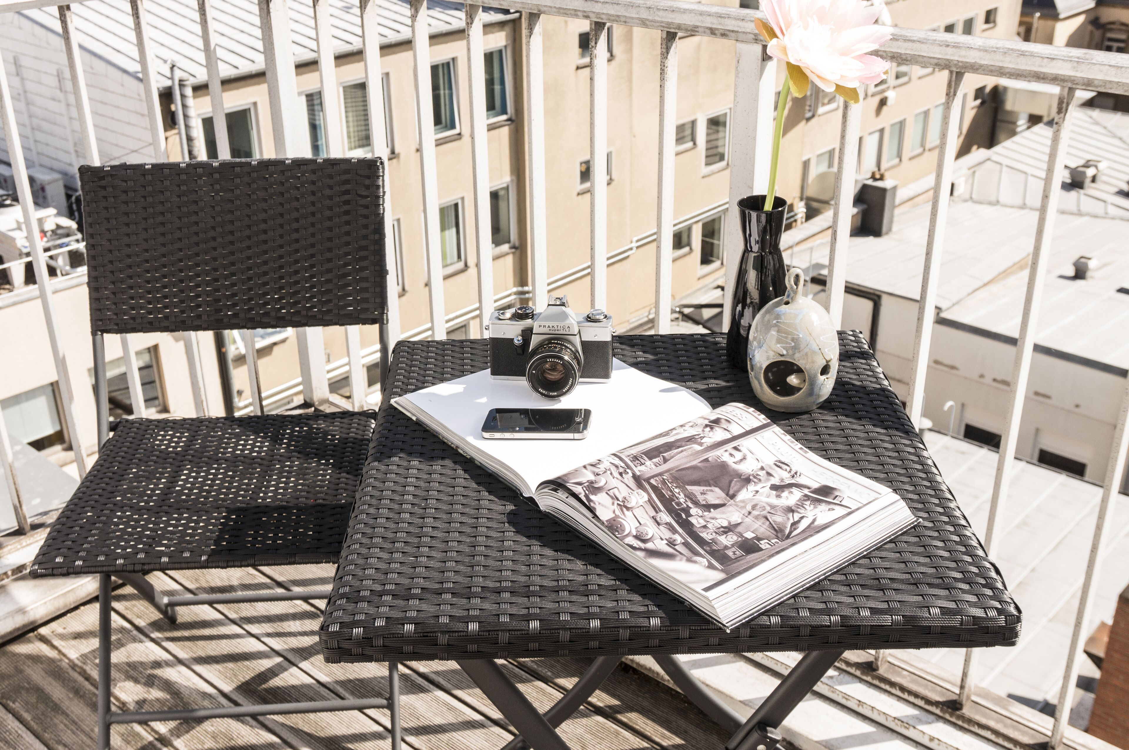A balcony with a wicker table, a camera, a smartphone, a vase with a flower, and a magazine.