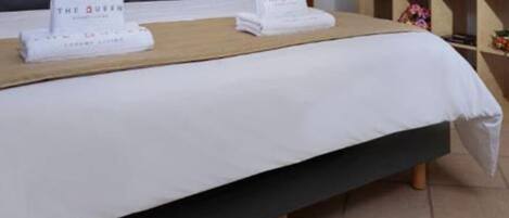 Una cama bien hecha con una colcha blanca y dos almohadas, cada una con un logotipo. Se observa un cabecero de madera, una mesita de noche con una lámpara, y una estantería con objetos decorativos.