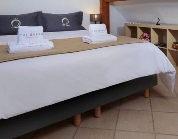 A neatly made bed with a white bedspread and two pillows, each with a logo. There is a wooden headboard, a bedside table with a lamp, and a shelf with decorative items.