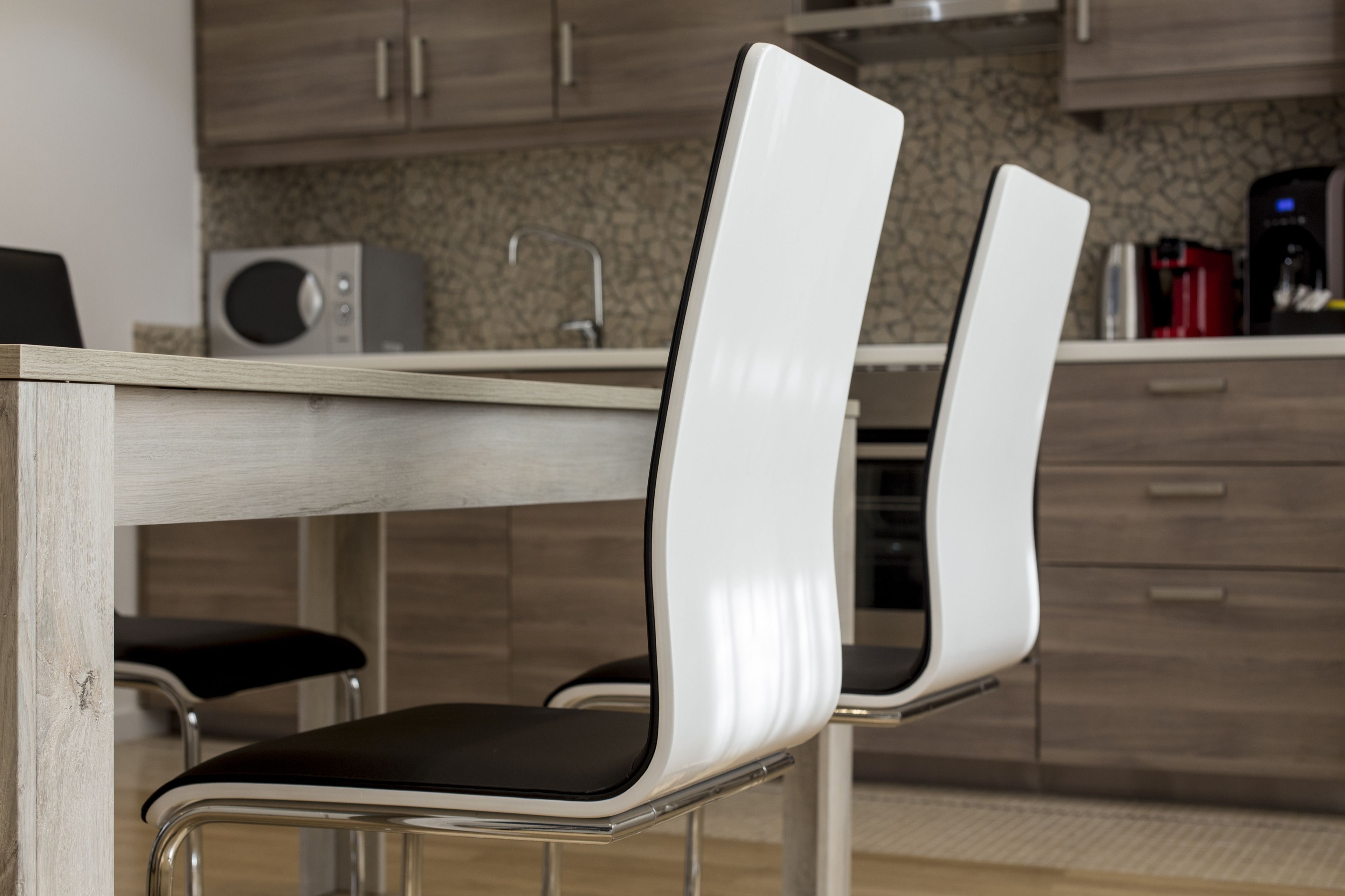 A modern kitchen with two white chairs with black cushions, a wooden table, and a coffee machine.