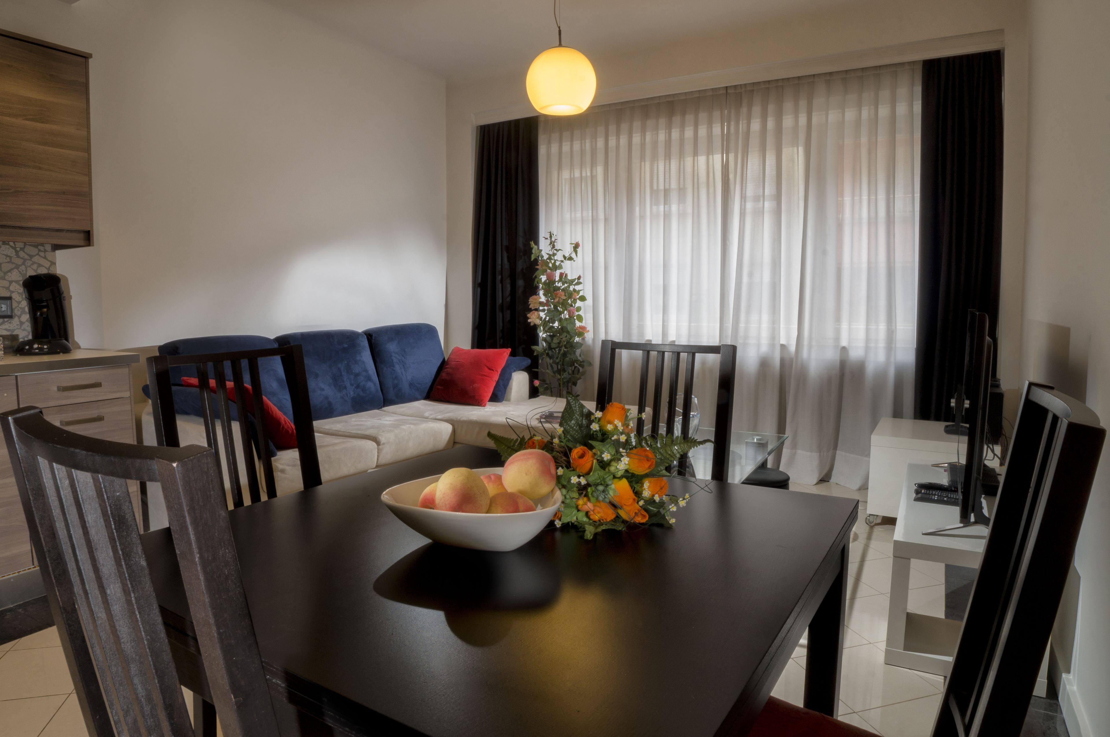Een moderne eetruimte met een donker houten tafel, een schaal fruit en een bloemstuk. Op de achtergrond staat een witte bank met blauwe en rode kussens, en een groot raam met transparante gordijnen.