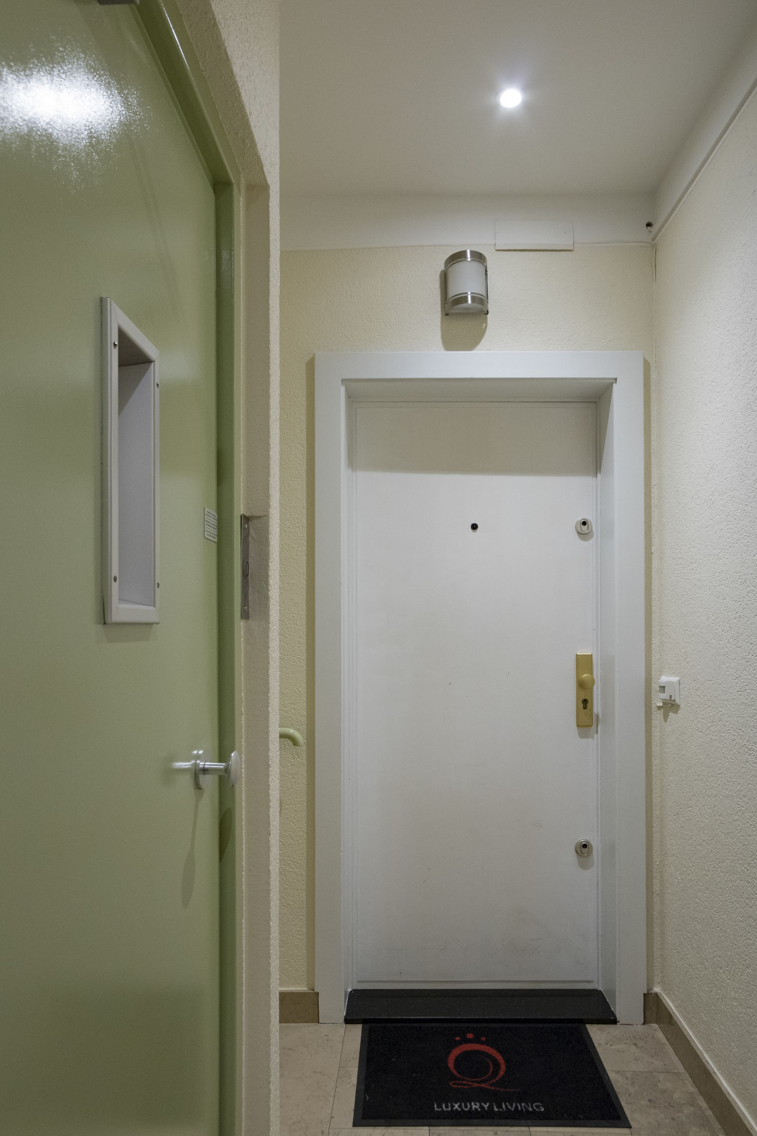 A hotel entrance with a green door, a white door with a security camera, and a black mat with "LUXURY LIVING" on it.