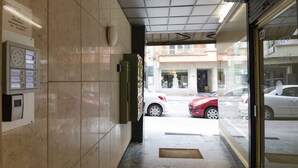 Hoteleingang mit einer gefliesten Wand, einem Gegensprechanlage und Blick auf die Straße mit parkenden Autos.