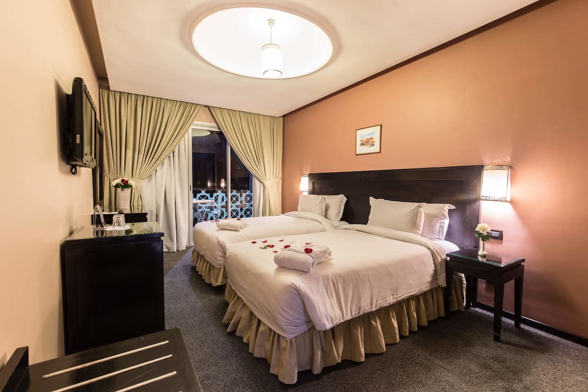 A hotel room with two beds, a round ceiling light, a bedside table with a lamp, and a view of a balcony.
