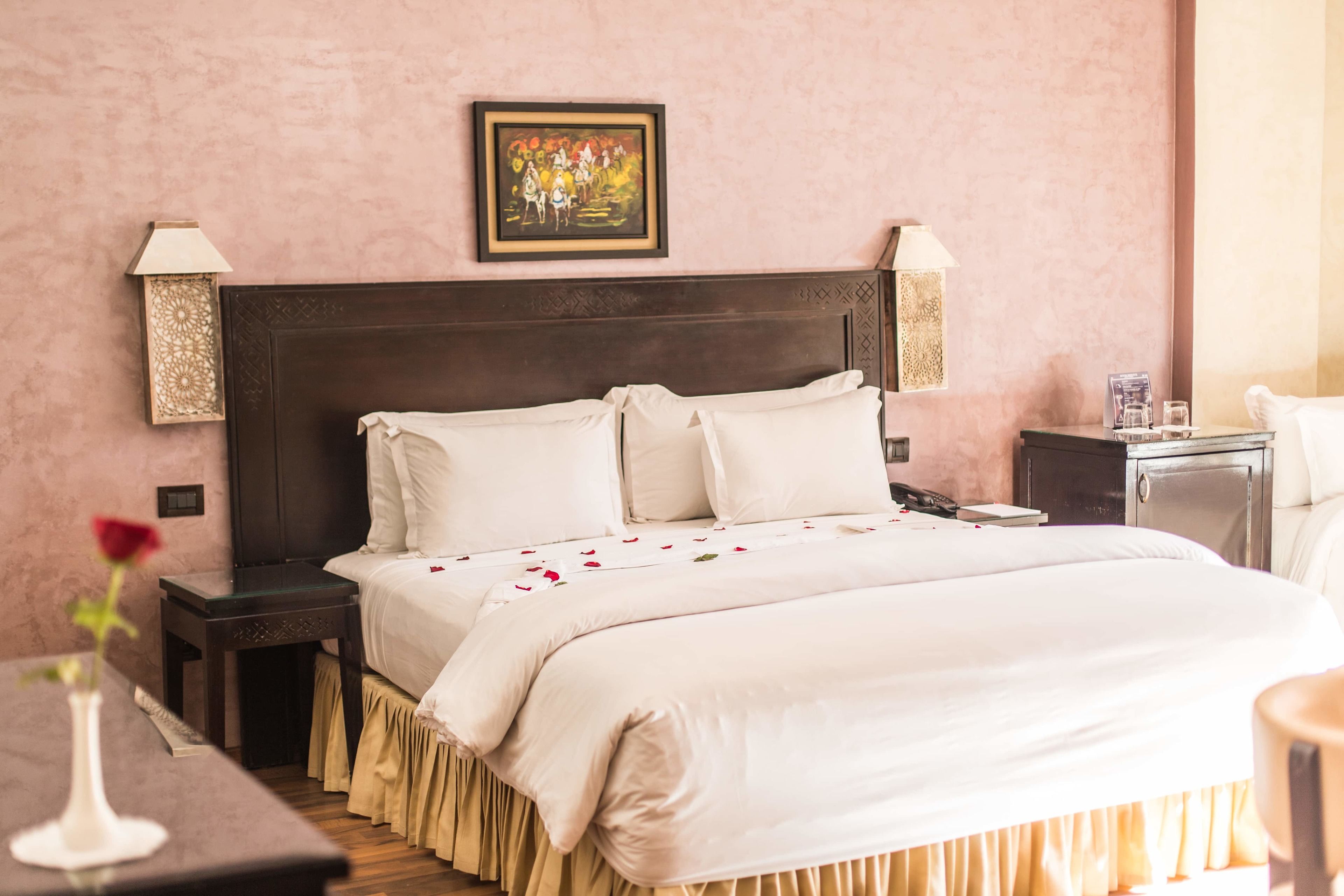 Una cama bien hecha con sábanas blancas y un cabecero decorativo, una mesita de noche con una lámpara, y una obra enmarcada sobre la cama.