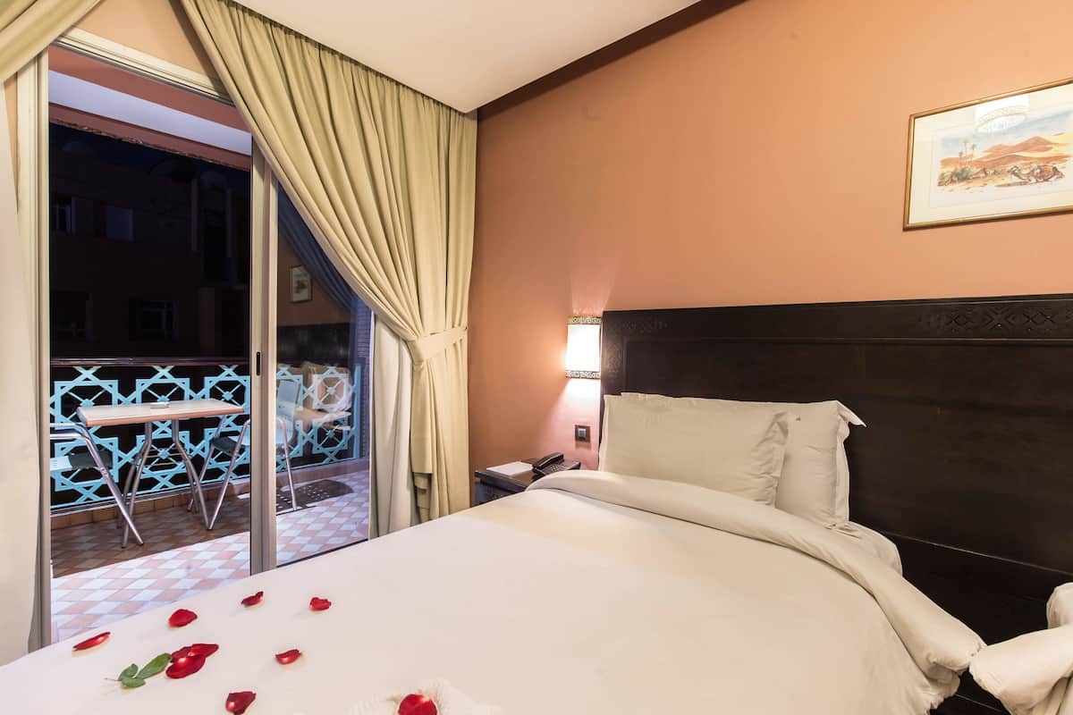 A hotel room with a bed, a nightstand, a lamp, and a view of an outdoor area with a table and chairs.