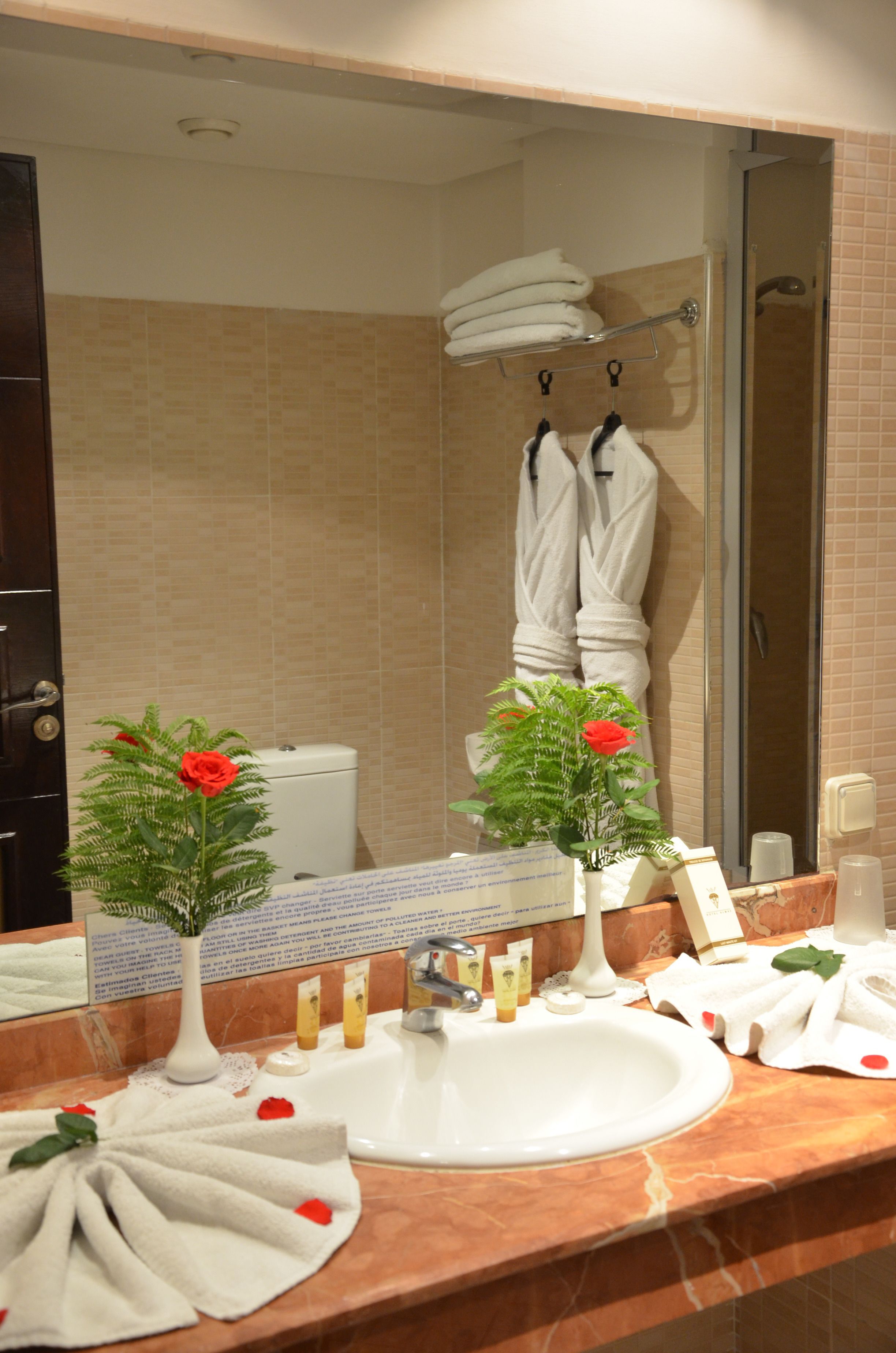Une salle de bain avec un lavabo, un miroir et deux vases contenant des fleurs artificielles. Des serviettes sont suspendues sur un porte-serviettes et une douche est visible au fond.