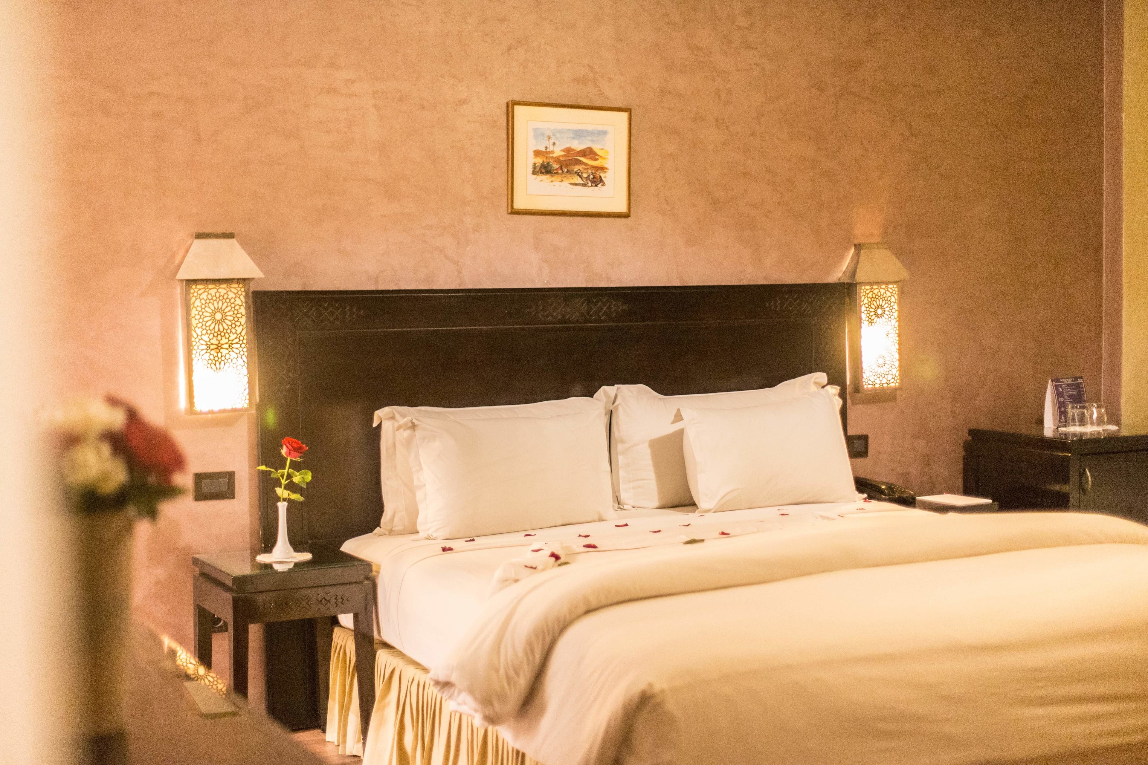 Una camera d'albergo con un letto grande, due comodini, un tavolino e un vaso con un fiore sul comodino.