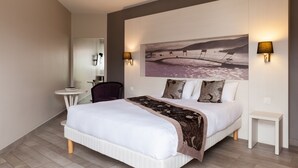 A modern bedroom with a large bed, a headboard with a painting, two bedside lamps, and a small table. - Charme Hôtel Montbéliard (Arbouans)