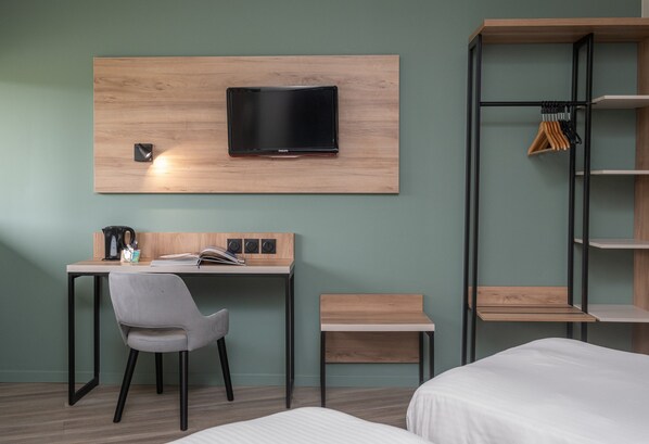A modern hotel room with a wooden desk, a grey chair, a bed, a TV mounted on the wall, and a wooden shelf unit. - Charme Hôtel Montbéliard (Arbouans)