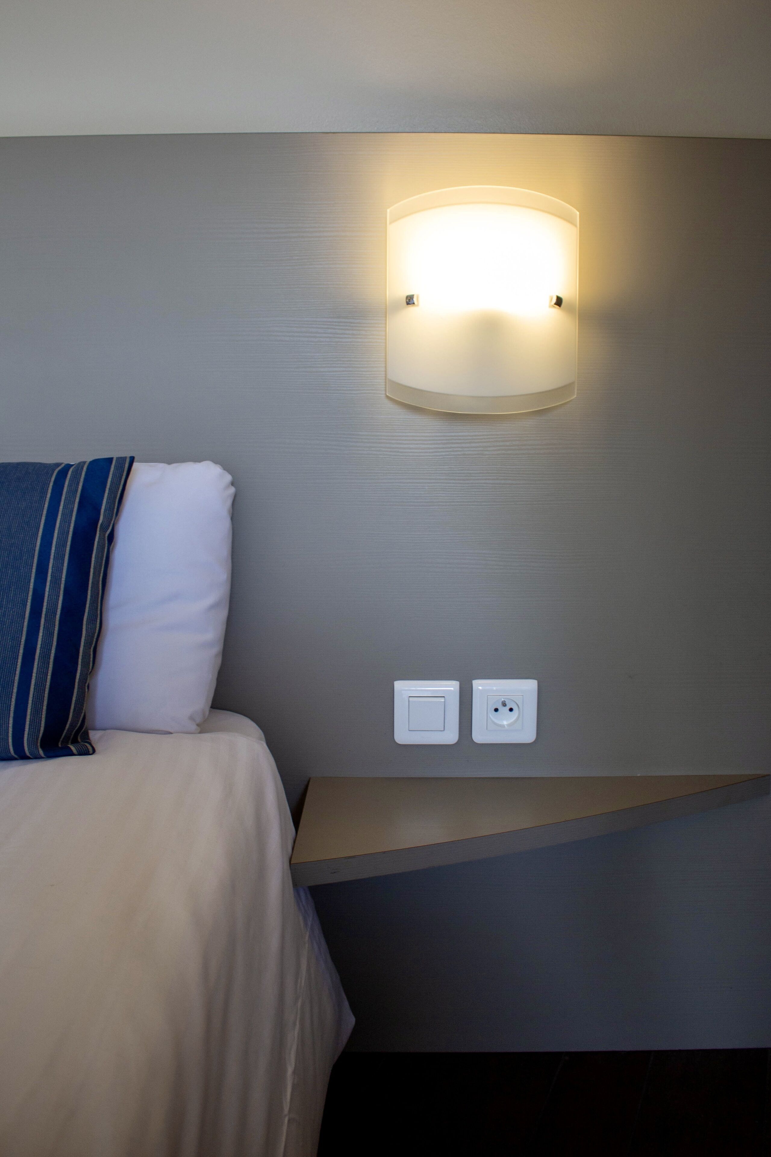 A neatly made bed with a striped pillow, a wall-mounted light fixture, and two electrical outlets on a bedside table.