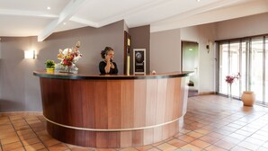 A reception desk with a woman standing behind it, a framed picture on the wall, and a large window with blinds. - Charme Hôtel Montbéliard (Arbouans)