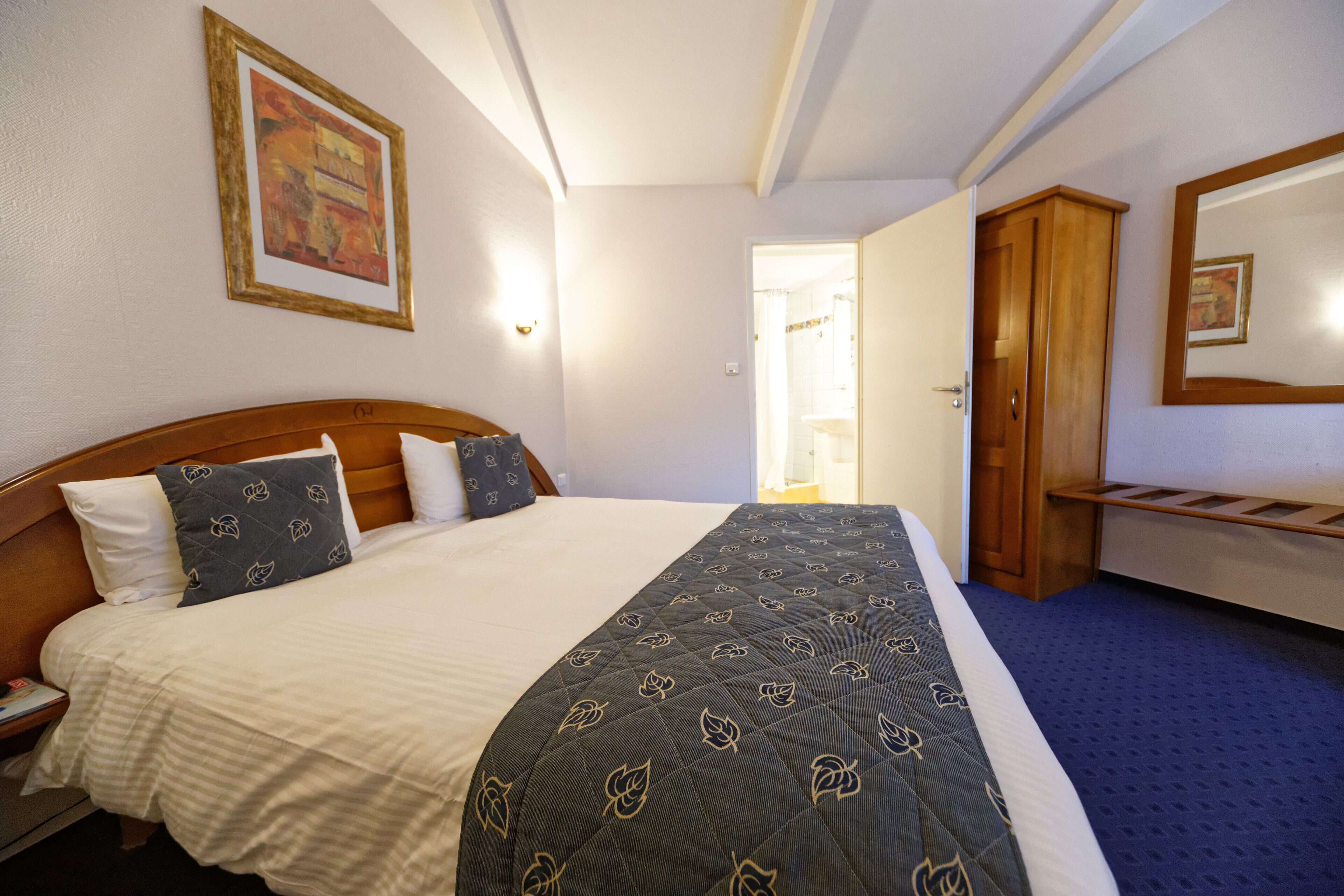 A hotel room with a large bed, a wooden wardrobe, a mirror, and a framed picture on the wall.