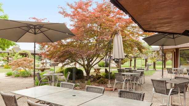 An outdoor patio area with multiple tables and chairs, surrounded by umbrellas and a garden with flowering trees. - Charme Hôtel Montbéliard (Arbouans)
