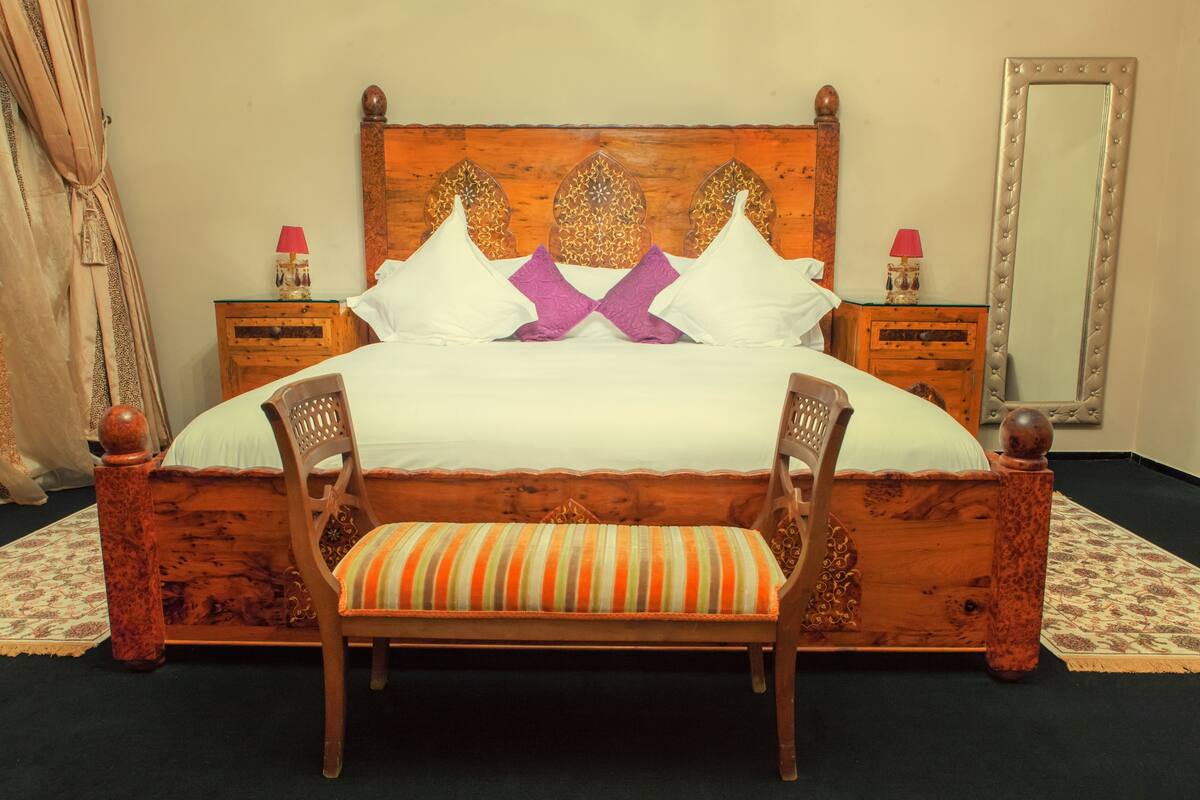 A wooden four-poster bed with white and purple pillows, a striped cushion, and two bedside tables with lamps.