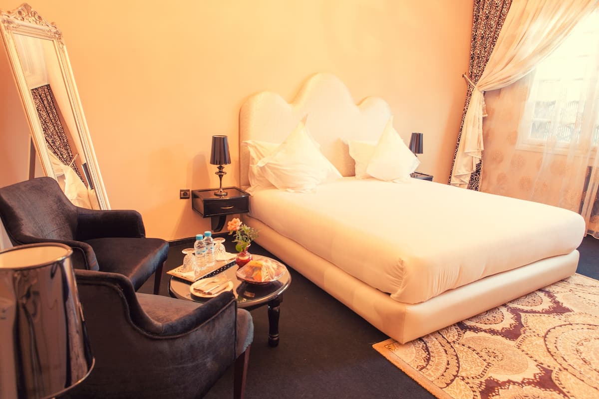 A hotel room with a large bed, a chair, a small table with a vase, and a mirror.