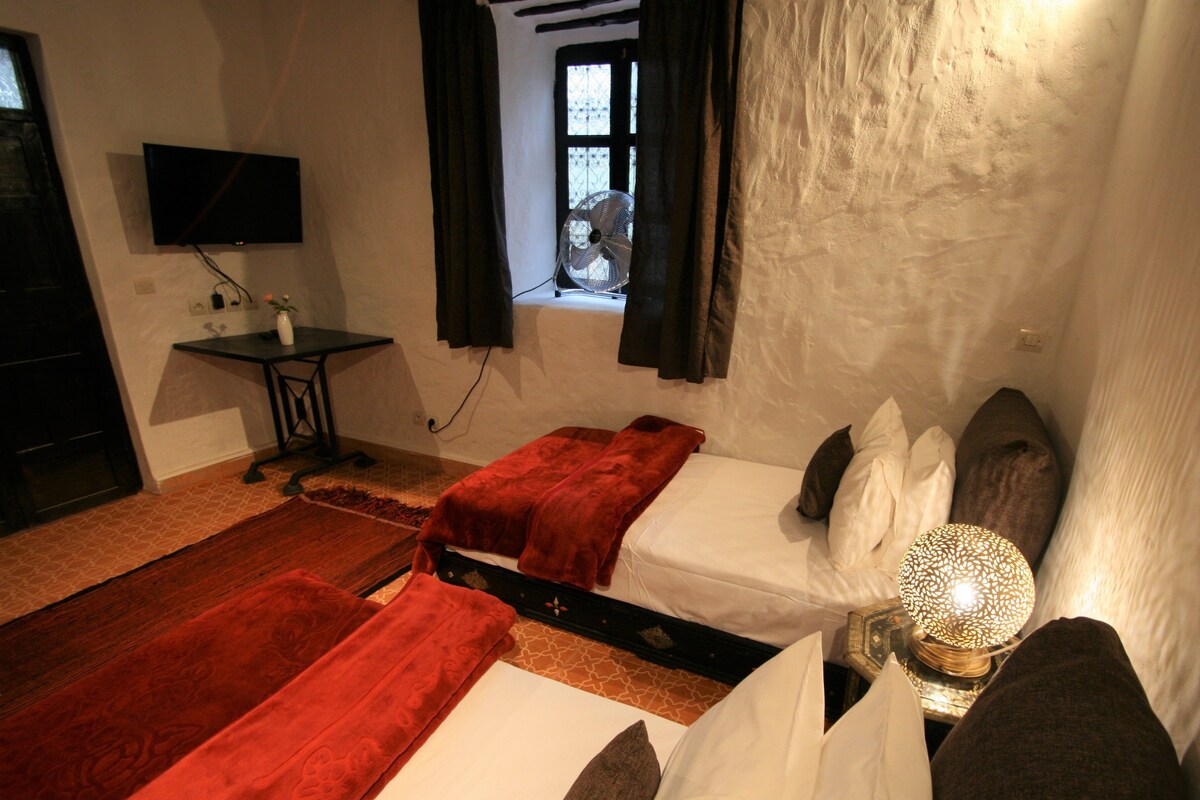 Ein Zimmer mit zwei Betten, einem Fernseher, einem Fenster mit Vorhängen, einem kleinen Tisch und einer Lampe.