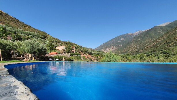 A clear blue pool surrounded by a stone deck, with a mountainous backdrop and a building with a red roof. - Kasbah Africa (Ouirgane)