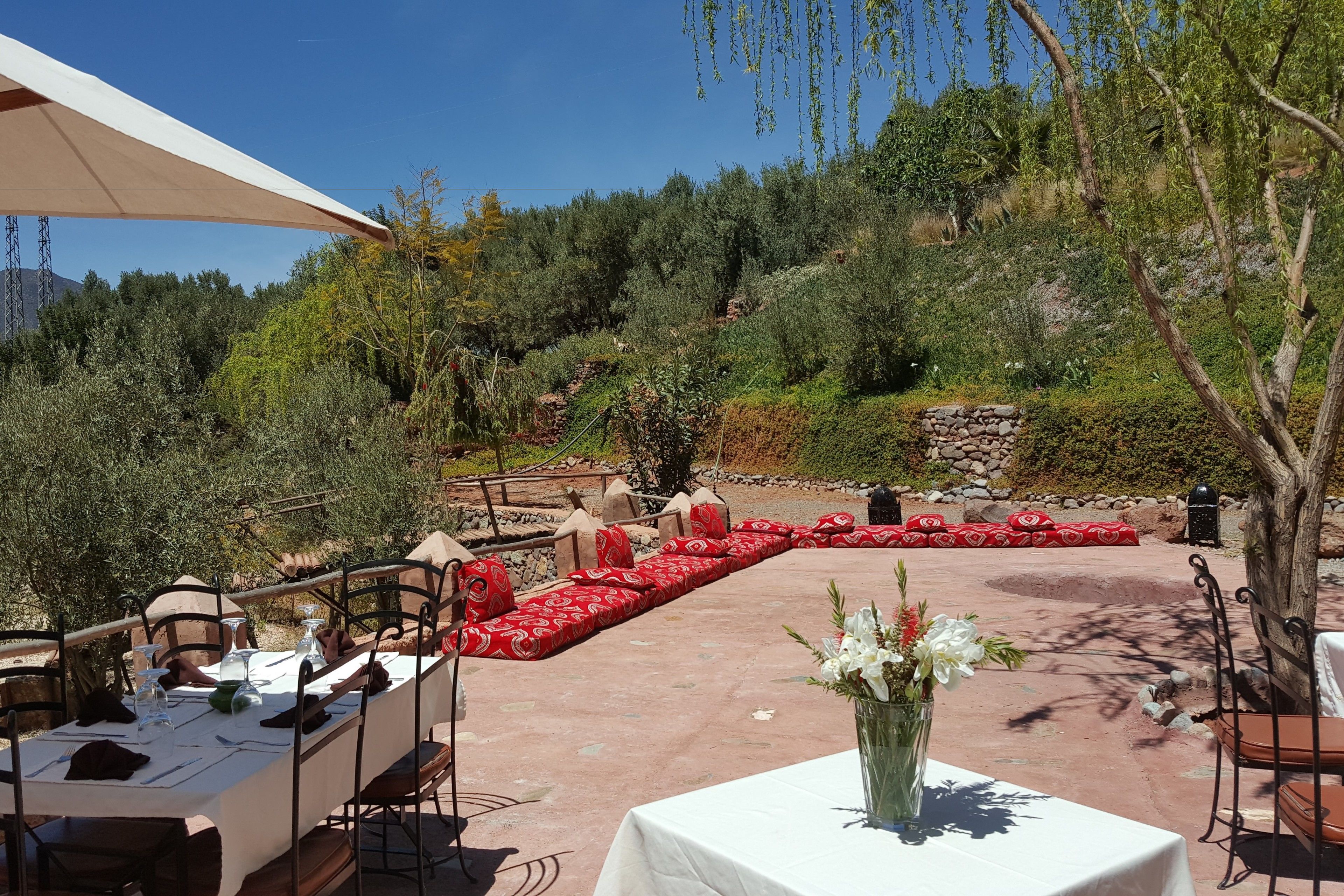 An outdoor dining area with tables and chairs, a stone wall, and a view of a hillside with vegetation.