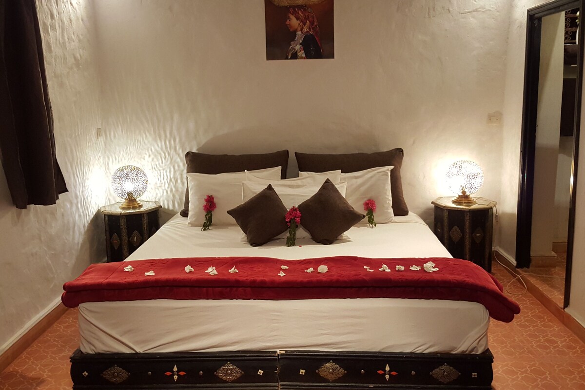 Un lit bien fait, avec une couverture rouge, deux lampes de chevet et des coussins décoratifs.