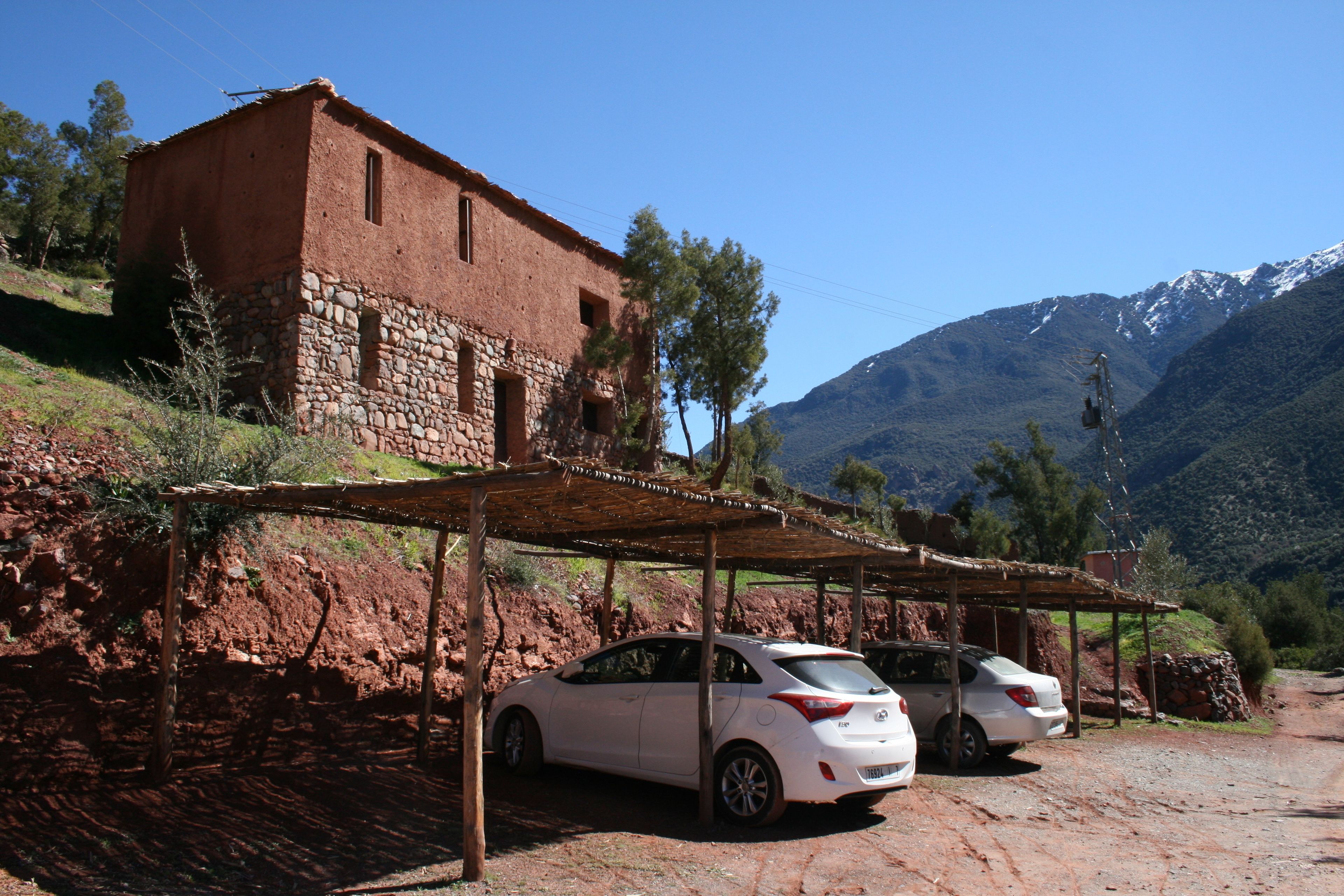 A rustic building with a stone base and a red upper structure, adjacent to a dirt road with parked cars.