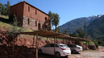 A rustic building with a stone base and a red upper structure, adjacent to a dirt road with parked cars.