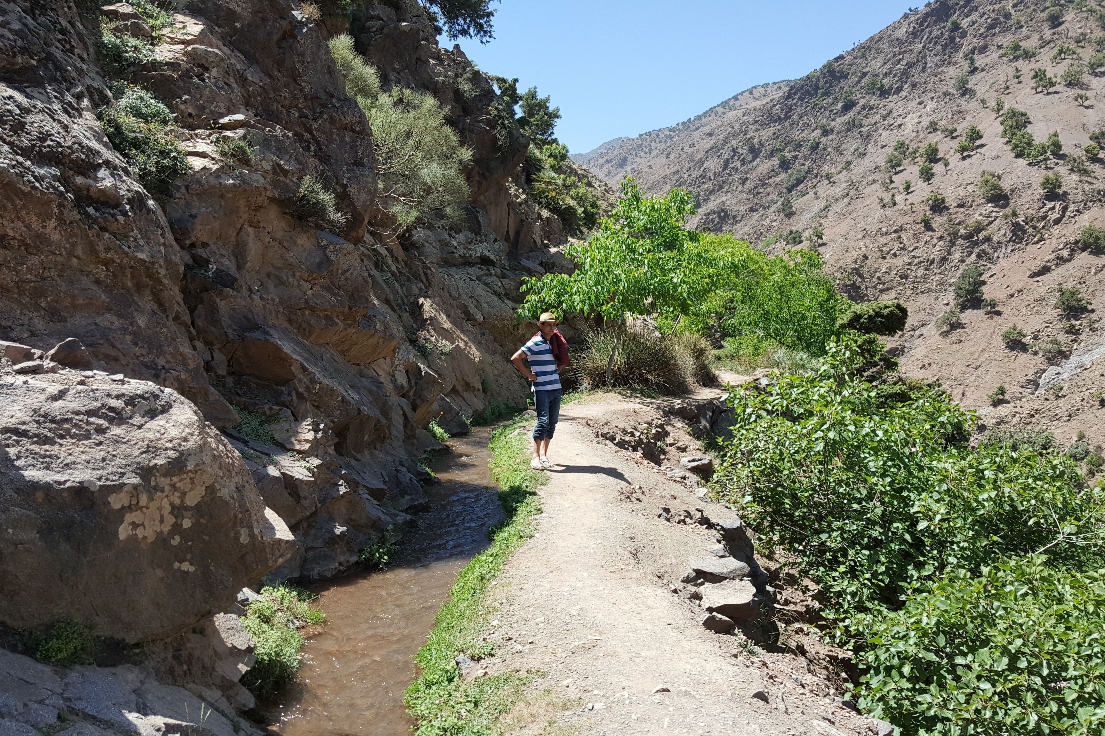A person walking on a narrow dirt path beside a rocky cliff with sparse vegetation.