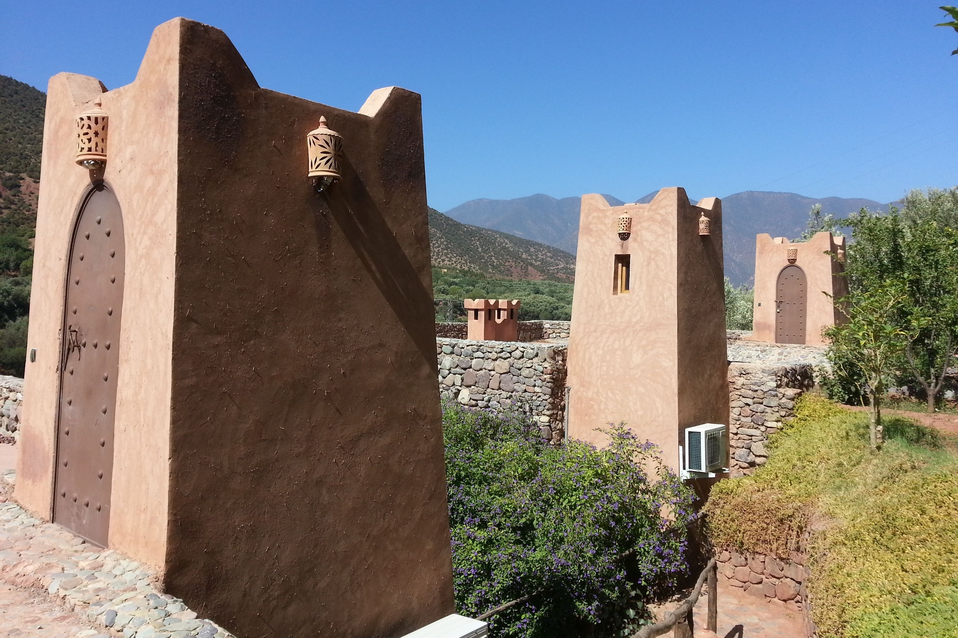 A traditional building with a large door and small windows, surrounded by greenery and mountains in the background.