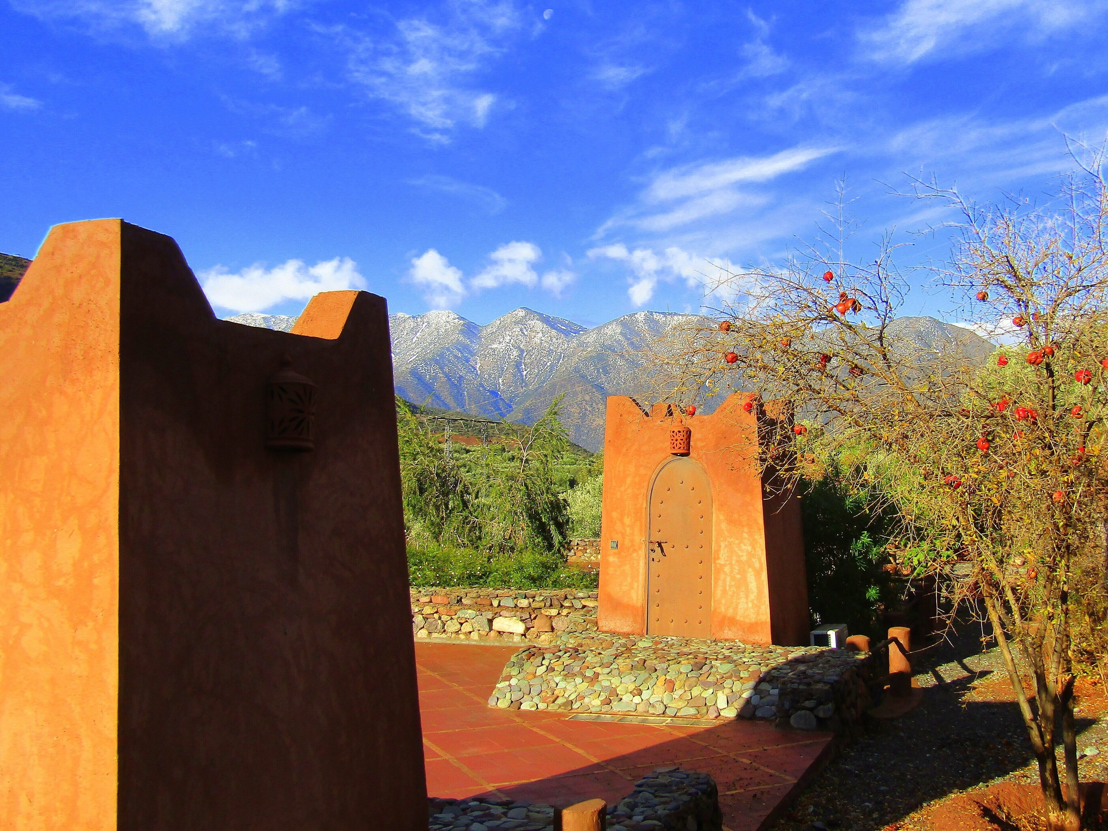A rustic building with a stone pathway leading to a wooden door, surrounded by mountains and a clear sky.