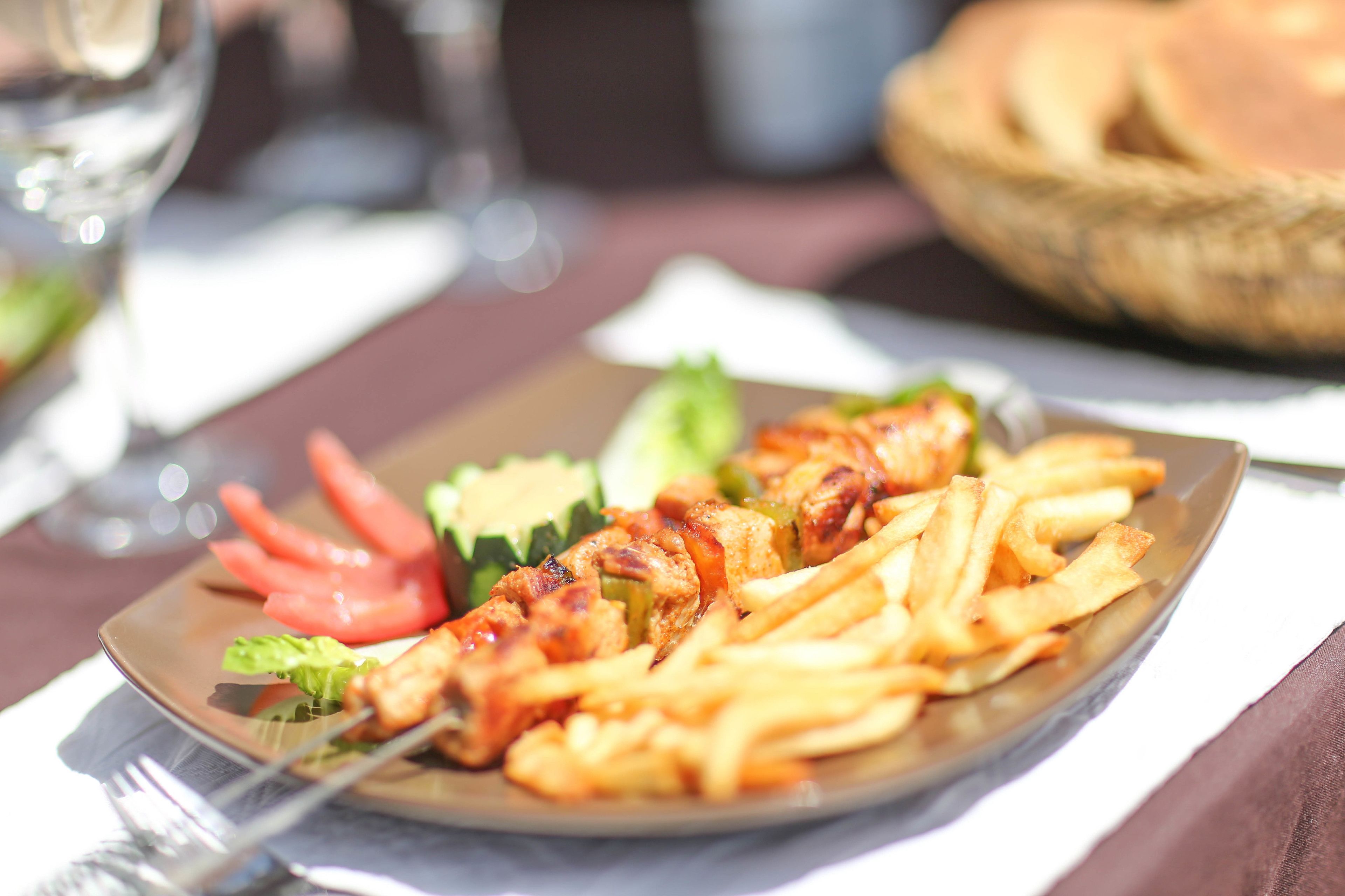 A plate of food with skewers and fries, a glass of water, and a basket of bread in the background.