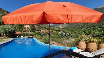 Una piscina con borde azul, rodeada de sillones de descanso y un paraguas naranja.
