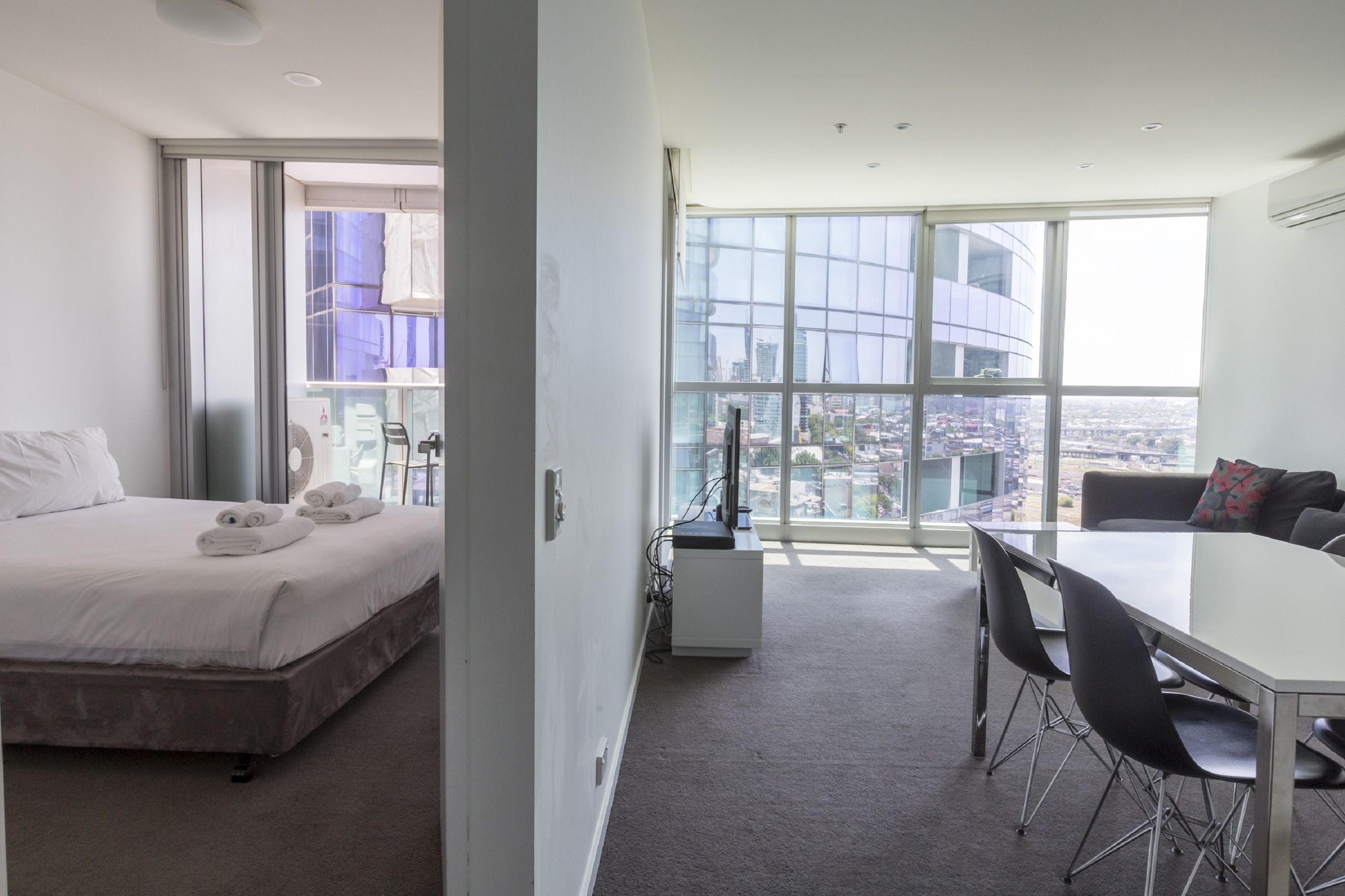 A modern hotel room with a large bed, a dining area, and a city view through floor-to-ceiling windows.