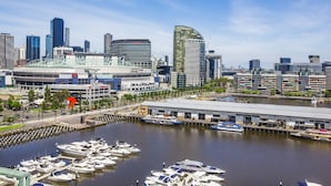A marina with boats docked, a modern city skyline in the background, and a large stadium-like structure with a curved roof. - Apartments Melbourne Domain - New Quay Docklands (Docklands)