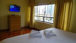 A hotel room with a bed, a wooden dresser, a television on the wall, and a window with curtains. - Suites Larco 656 (Lima)