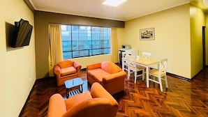 A living room with a large window, a dining table, and two orange sofas. - Suites Larco 656 (Lima)