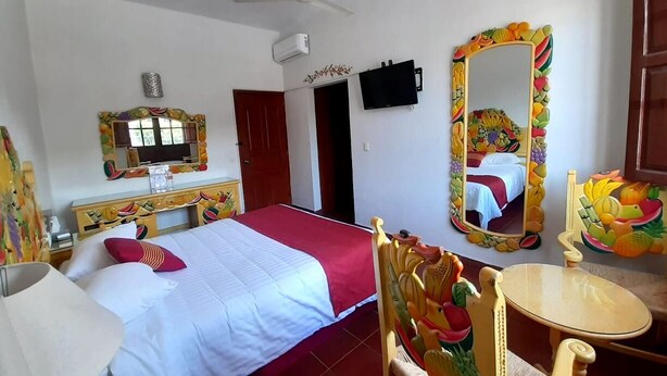 A hotel room with a bed, a TV, a ceiling fan, a mirror with a fruit frame, and a small table.