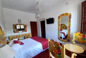Quarto de hotel com cama, televisão, ventilador de teto, espelho com moldura de frutas e uma pequena mesa.