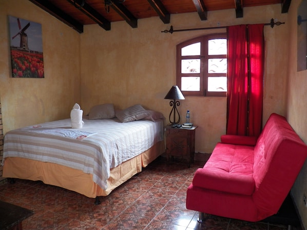 A bedroom with a bed, a red chair, a nightstand with a lamp, a window with red curtains, and a wall painting. - Hotel Ciudad Condal (Antigua Guatemala)