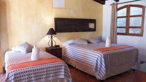 A room with two beds, each with a patterned bedspread and pillows, a nightstand with a lamp, and a window with curtains. - Hotel Ciudad Condal (Antigua Guatemala)