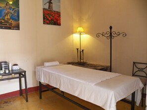 Quarto pequeno e simples com uma cama de solteiro, uma mesa com produtos de higiene pessoal, um abajur e um pôster de moinho de vento na parede.