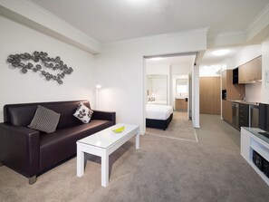 Ein modernes Hotelzimmer mit einem dunkelbraunen Sofa, einem weißen Couchtisch, einem Bett mit weißer Matratze, einem Badezimmer mit Waschbecken und Spiegel sowie einer Küchenzeile mit Mikrowelle und Schränken.