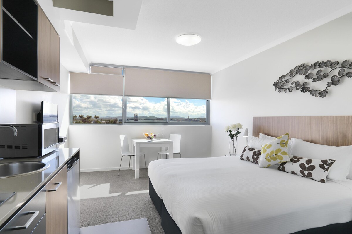 A modern hotel room with a bed, a desk, a chair, a kitchen area, and a view of the outdoors.
