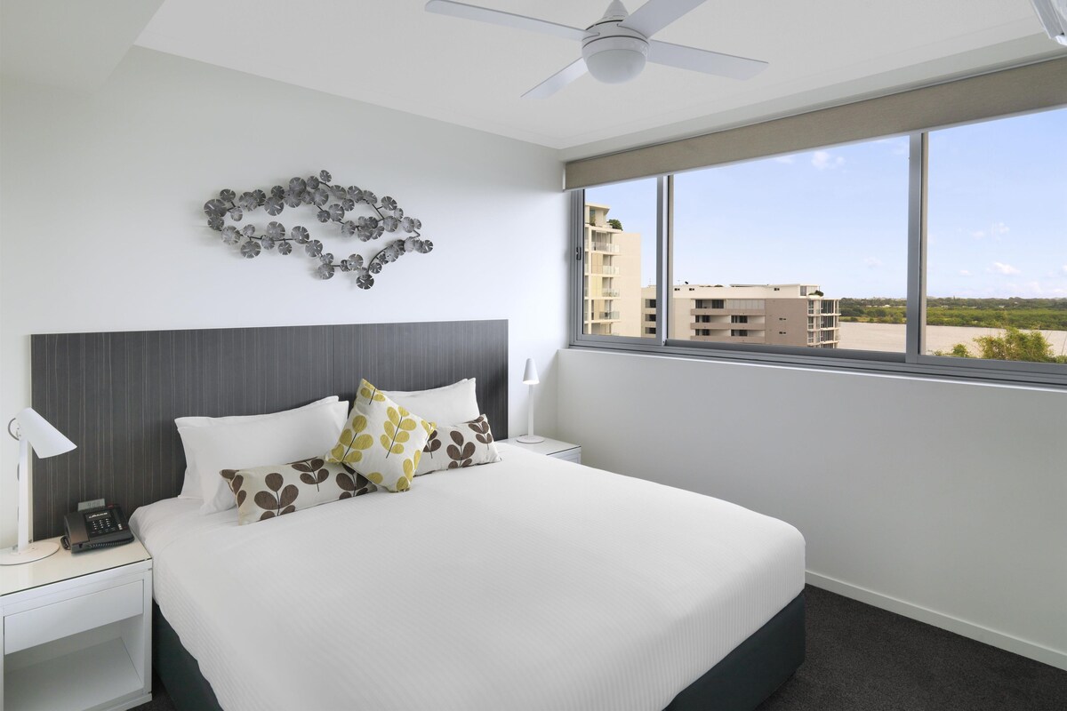 A hotel room with a bed, a nightstand, a ceiling fan, a window with a view of buildings, and a wall decoration.
