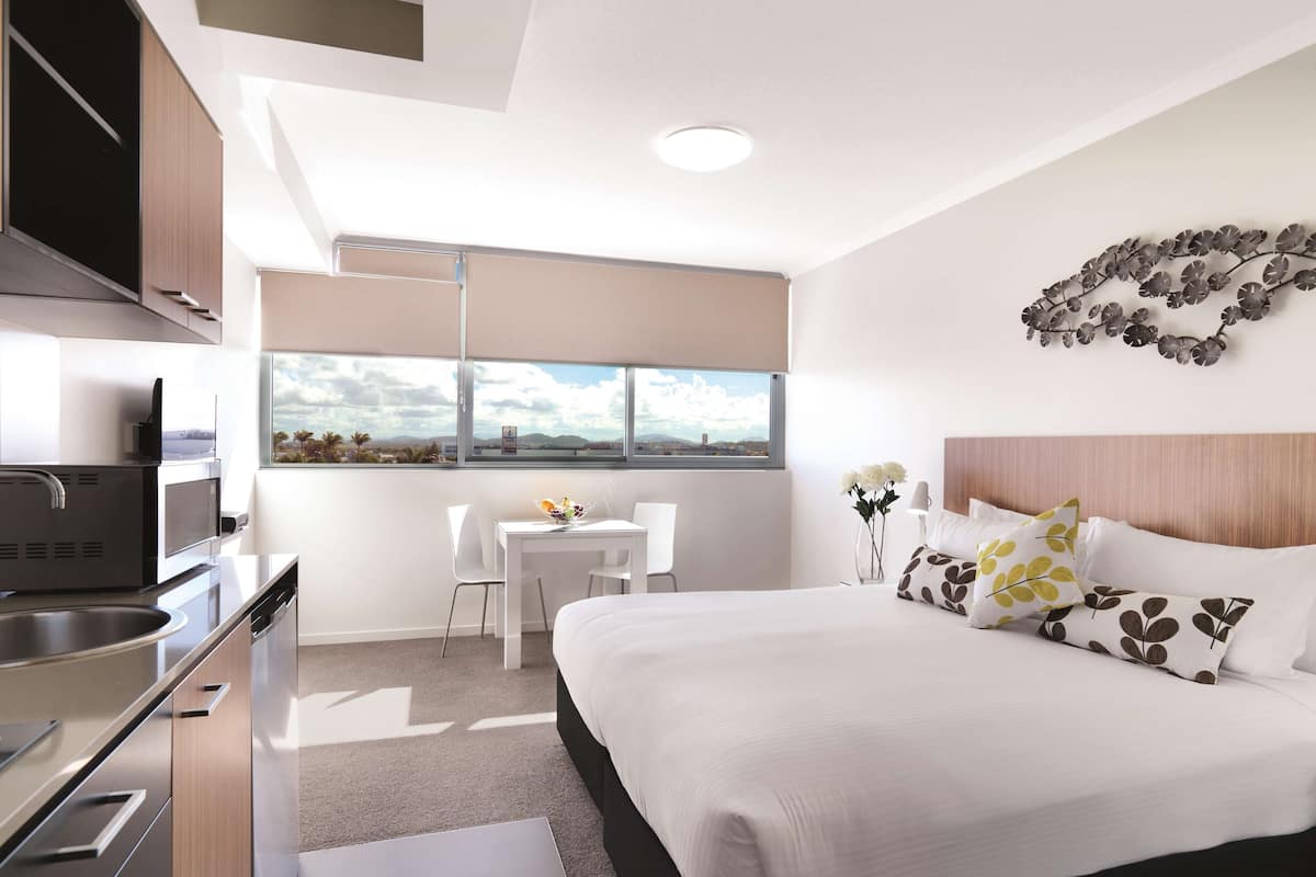 A modern hotel room with a bed, a desk, a chair, a view of the city, and a kitchenette.
