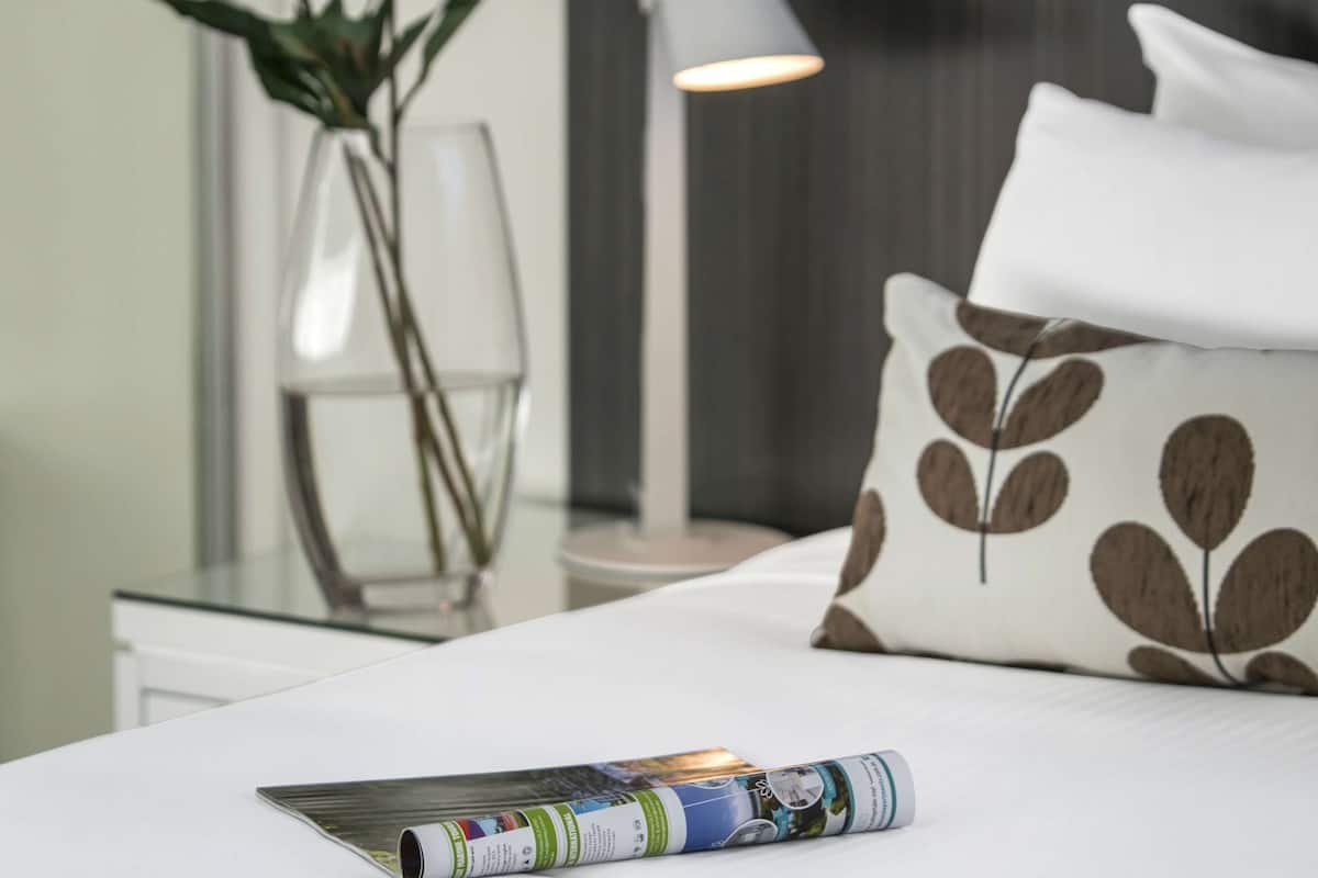 A neatly made bed with a pillow featuring a leaf pattern, a clear vase with white flowers, and a magazine on the bed.