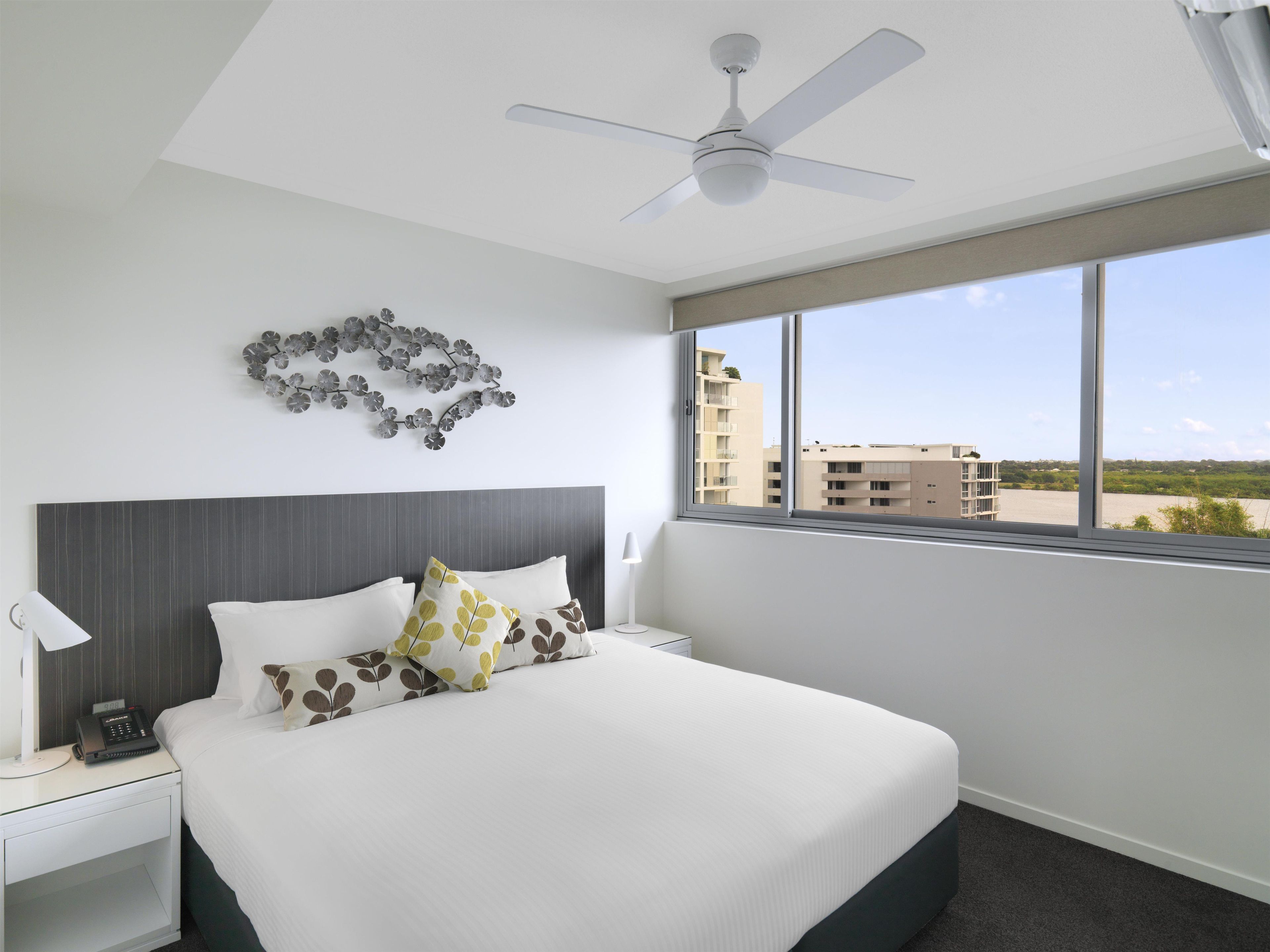 Quarto de hotel com cama, mesa de cabeceira, ventilador de teto, janela com vista para edifícios e um enfeite na parede.
