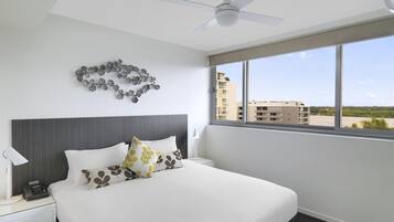 Quarto de hotel com cama, mesa de cabeceira, ventilador de teto, janela com vista para edifícios e um enfeite na parede.