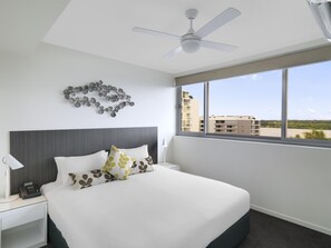 A hotel room with a bed, a nightstand, a ceiling fan, a window with a view of buildings, and a wall decoration. - Oaks Mackay Rivermarque Hotel (Mackay)