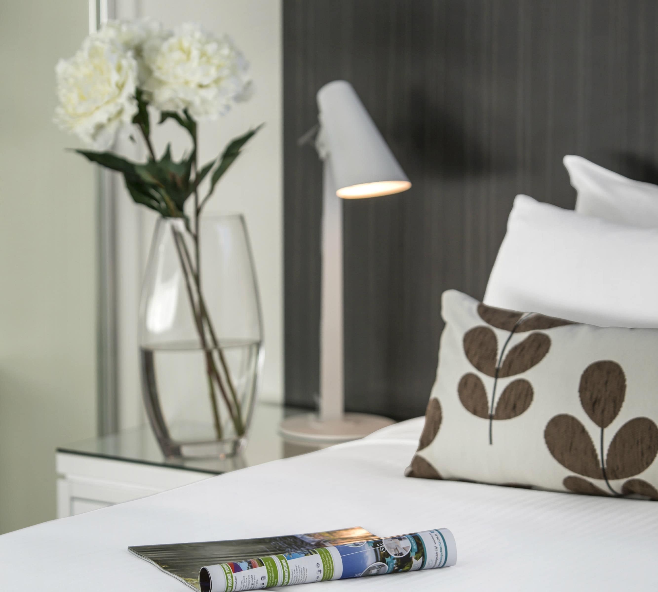 Una cama bien hecha con un cojín, una revista, un florero con flores y una lámpara.