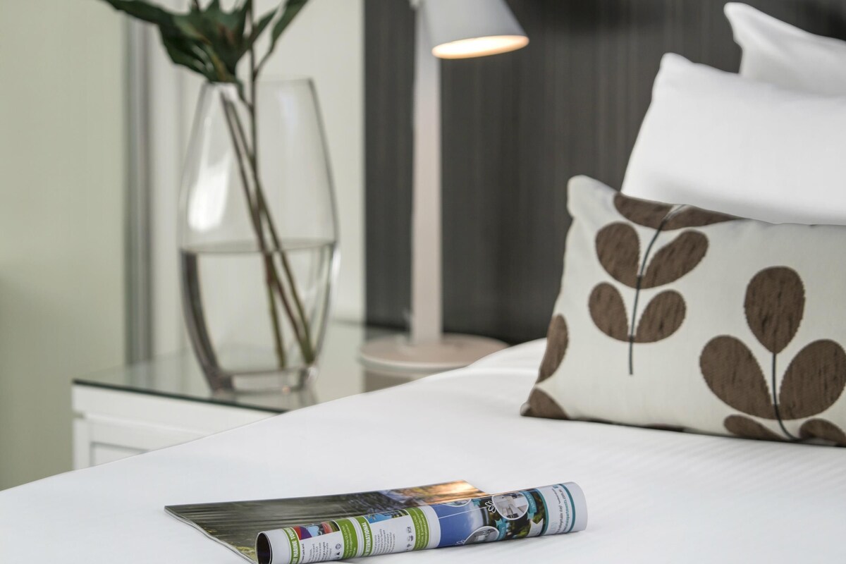 A neatly made bed with a pillow, a magazine, a vase with flowers, and a lamp.